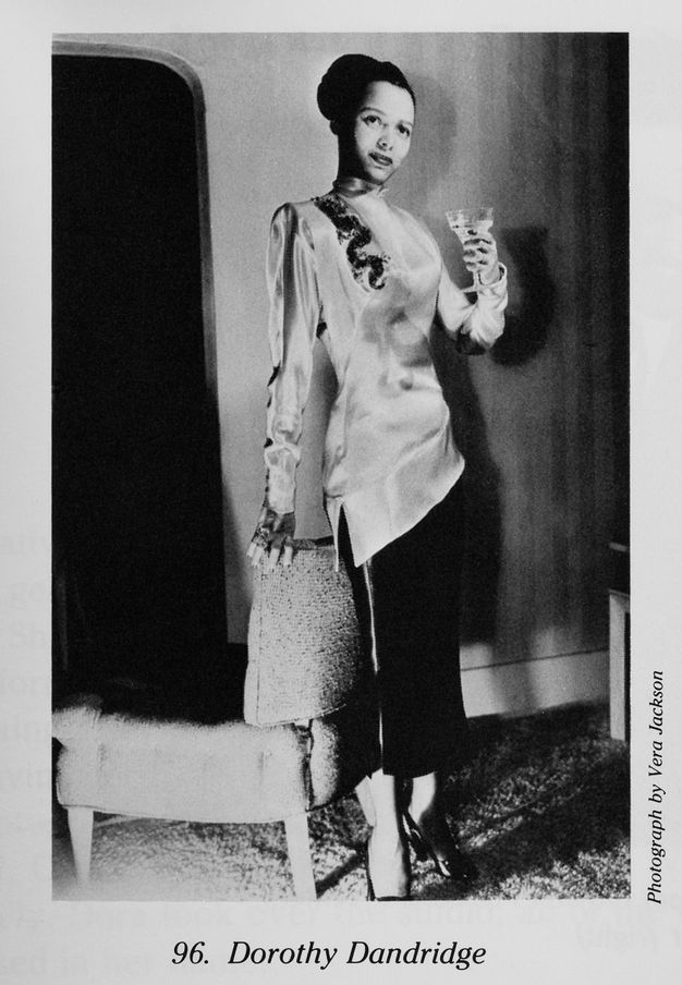 A black-and-white photograph depicting a standing figure as she raises a stemmed glass in one hand and touches the back of an upholstered chair with the other. The spotlighting and shadows, along with her fancy evening attire, convey a mood of importance and drama.