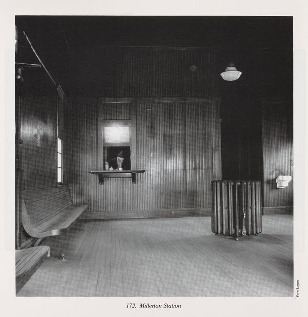 Black-and-white photograph of a dated, near-empty waiting room in a train station, full of wood paneling and accent lighting. In the back wall behind a ticket window, a cashier is framed by a bright light above his counter. A caption reads “172. Millerton Station.”