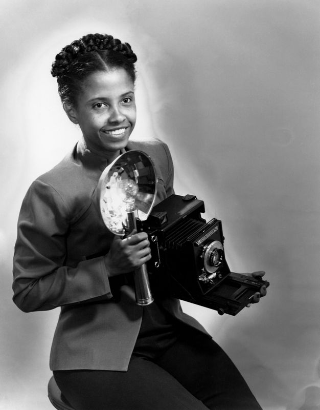 A black-and-white, half-length photograph portrait of a seated photographer, who smiles and looks off into the distance as she holds her large camera and its flash close to her chest. Her hair is neatly groomed with a short part down the middle.