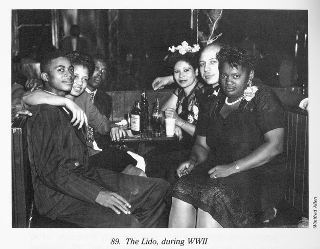 A black-and-white photograph of six well-dressed men and women seated around a round table full of bottles and drinking glasses. The ambiance is enhanced with low lighting in the night club.