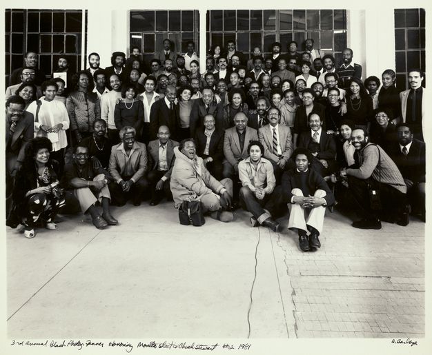 A black-and-white portrait of a large group posing and smiling for the camera, those in the front kneeling or seated. People stand in front of gridded windows that reveal an evening setting. A handwritten caption details the date and occasion of the gathering. A signature appears on the bottom right corner.