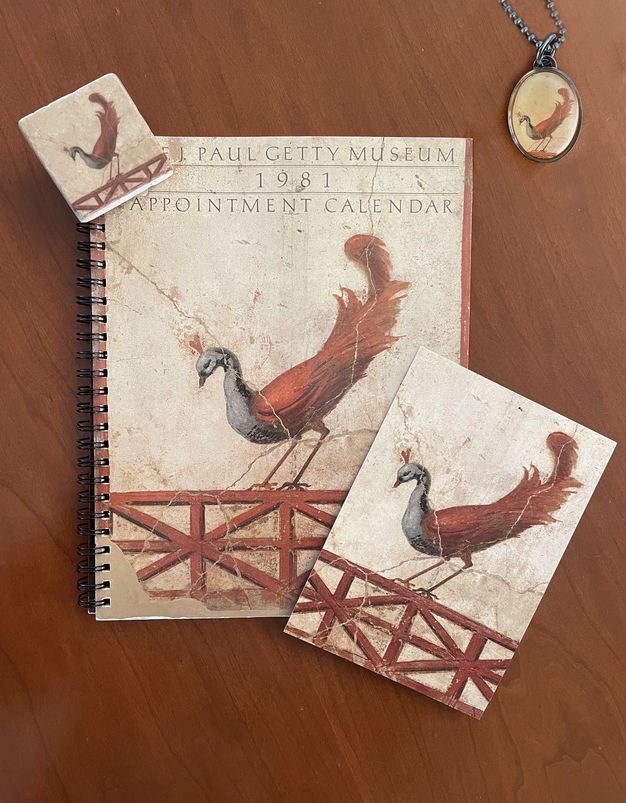 An array of printed and bound objects and the pendant of a necklace on a chain, all featuring a reproduction of a blue-and-red peacock in various cropped views.