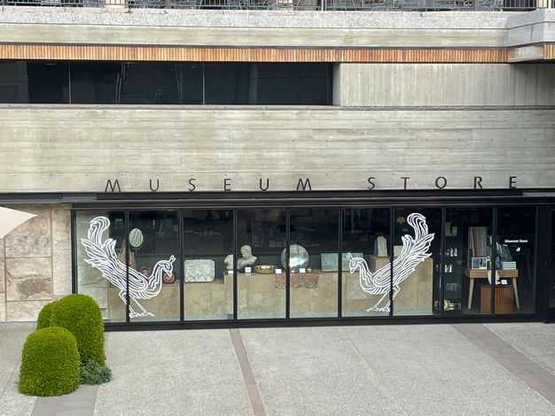 Viewed from in an outdoor courtyard, a storefront whose full-length windows feature white-stenciled silhouettes of two facing peacocks from floor to ceiling.