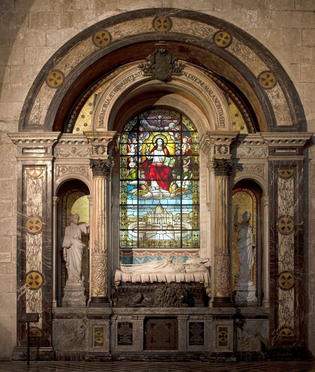 A multipart funerary monument within an arched space. At the center, a marble figure reclines in front of a stained-glass window Flanked by ornate columns holding up a second, smaller arch. A pair of niches house statues on either side.