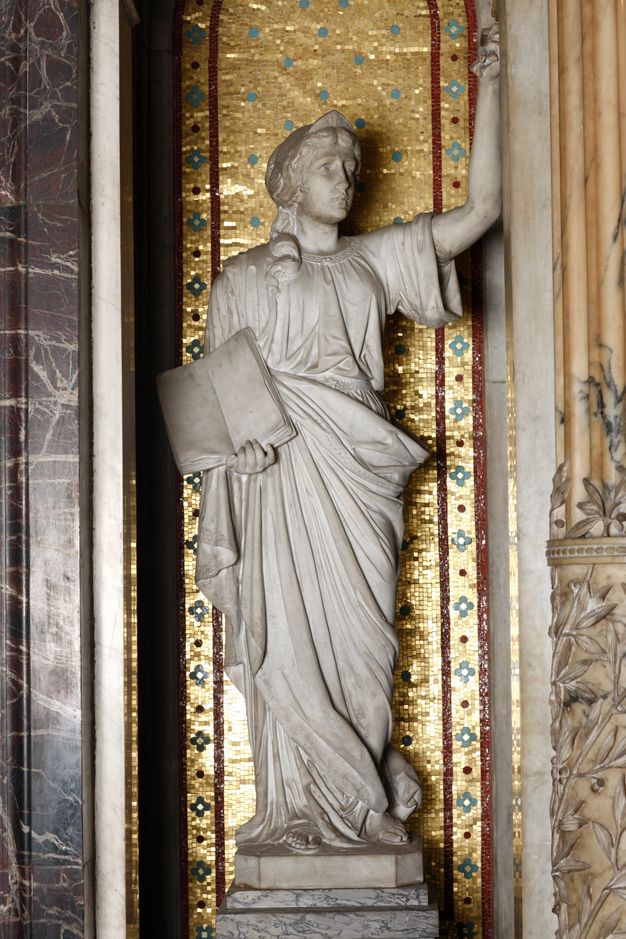 A sculpture in a mosaic-lined niche depicts a draped female figure with one hand raised and the other holding an open book.