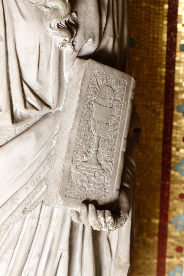 Detail of a marble sculpture in which a hand holds a closed book. On the book cover, religious symbols and clouds are set off by a textured background.