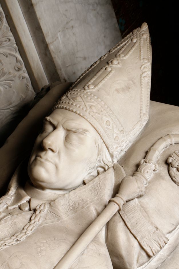 Close-up of a funerary sculpture depicting a man’s head and shoulders in episcopal attire. He lies on a pillow with his eyes closed. A staff with a curved head rests over his shoulder.