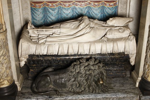 A funerary sculpture depicting an archbishop lying supine on draped fabric with his head and feet oriented to the surrounding columns. A bronze lion crouches below him.