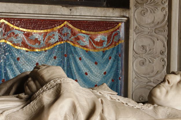 A close-up of the mosaic wall behind a supine sculpture of an archbishop. The mosaic imitates a dotted drapery.