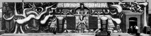 A black-and-white photograph of a long mural located on a rooftop showing the bottom of a pre-Columbian pyramid, surrounded by giant jungle vines. At center, a figure is bound to a wooden crucifix surmounted by an eagle, and, at right, two men, one in broad-brimmed hat, the other in a cloth cap with ear flaps, hold rifles at the ready.