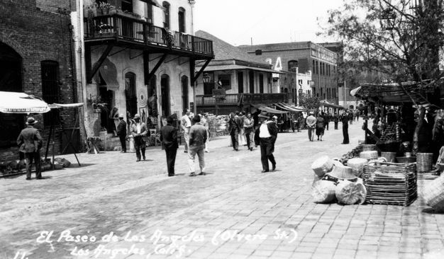 A black-and-white photograph shows a cobbled street with pedestrians strolling along turn-of-the-century brick buildings and awning-covered stalls selling woven baskets and other wares. White handwritten lettering in the bottom left gives the name of the street.
