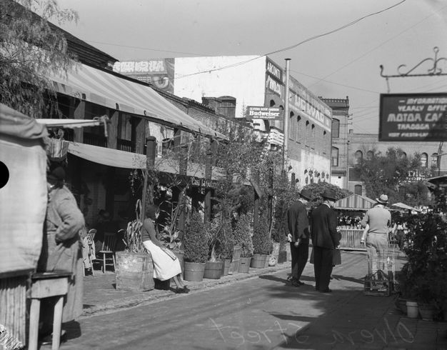 A black-and-white photograph of a street lined with brick buildings and dotted with pedestrians. Part of a mural can be seen on the second story of a building.