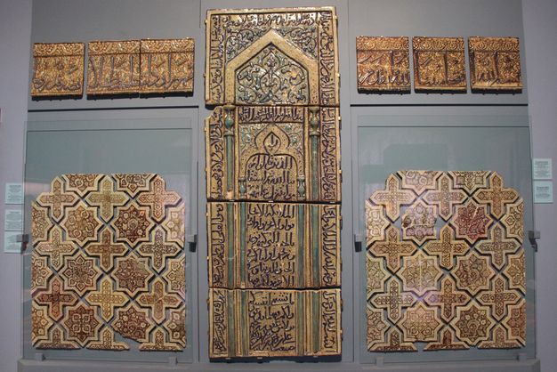 Groups of luster tiles mounted on a museum wall, including a tall middle panel, and smaller square panels with stars, crosses, and rectangles decorated with Arabic script to the upper and lower left and right.