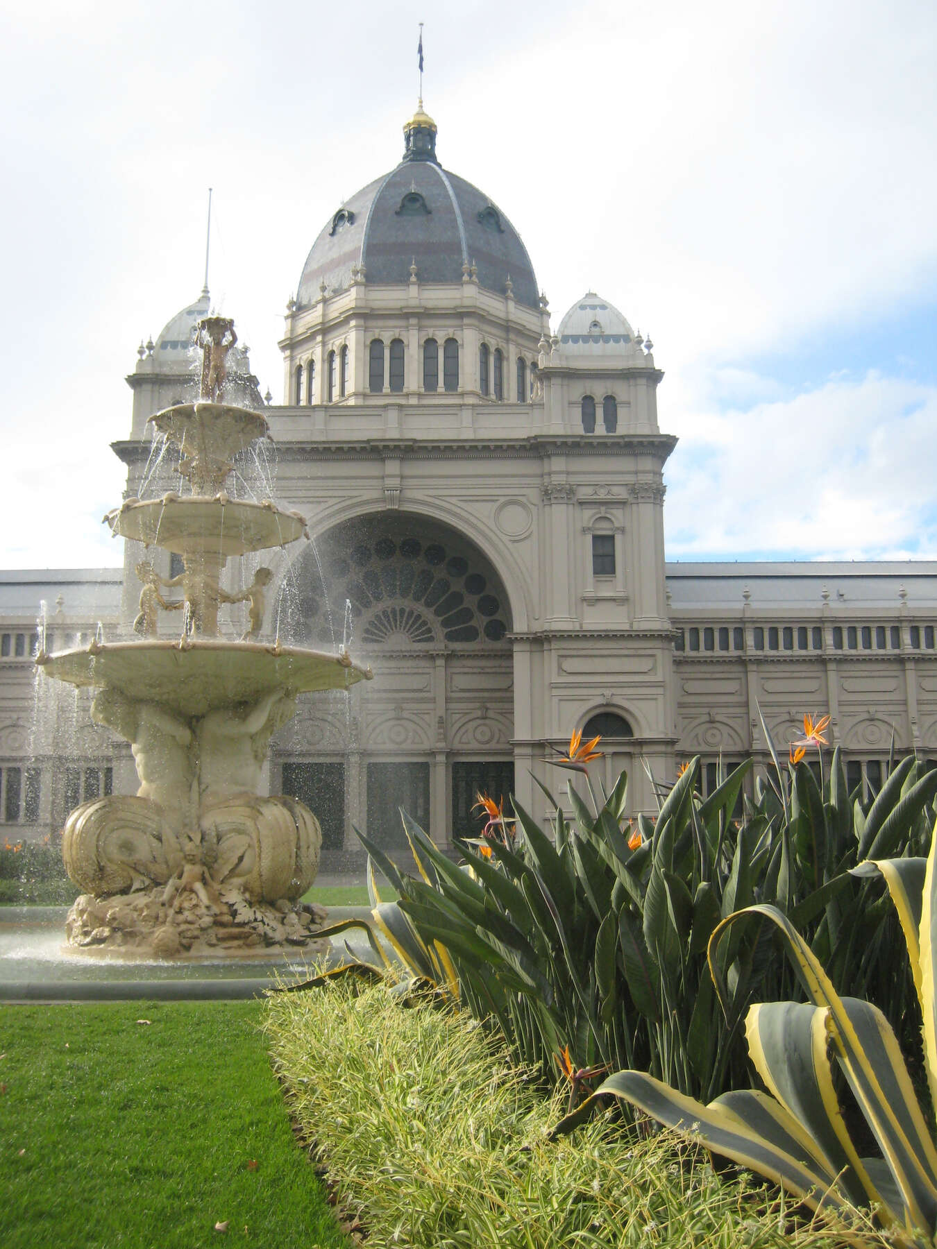 Sunny view of a palace and in front, a large garden filled with birds of paradise and a three-tier fountain with decorative putti.