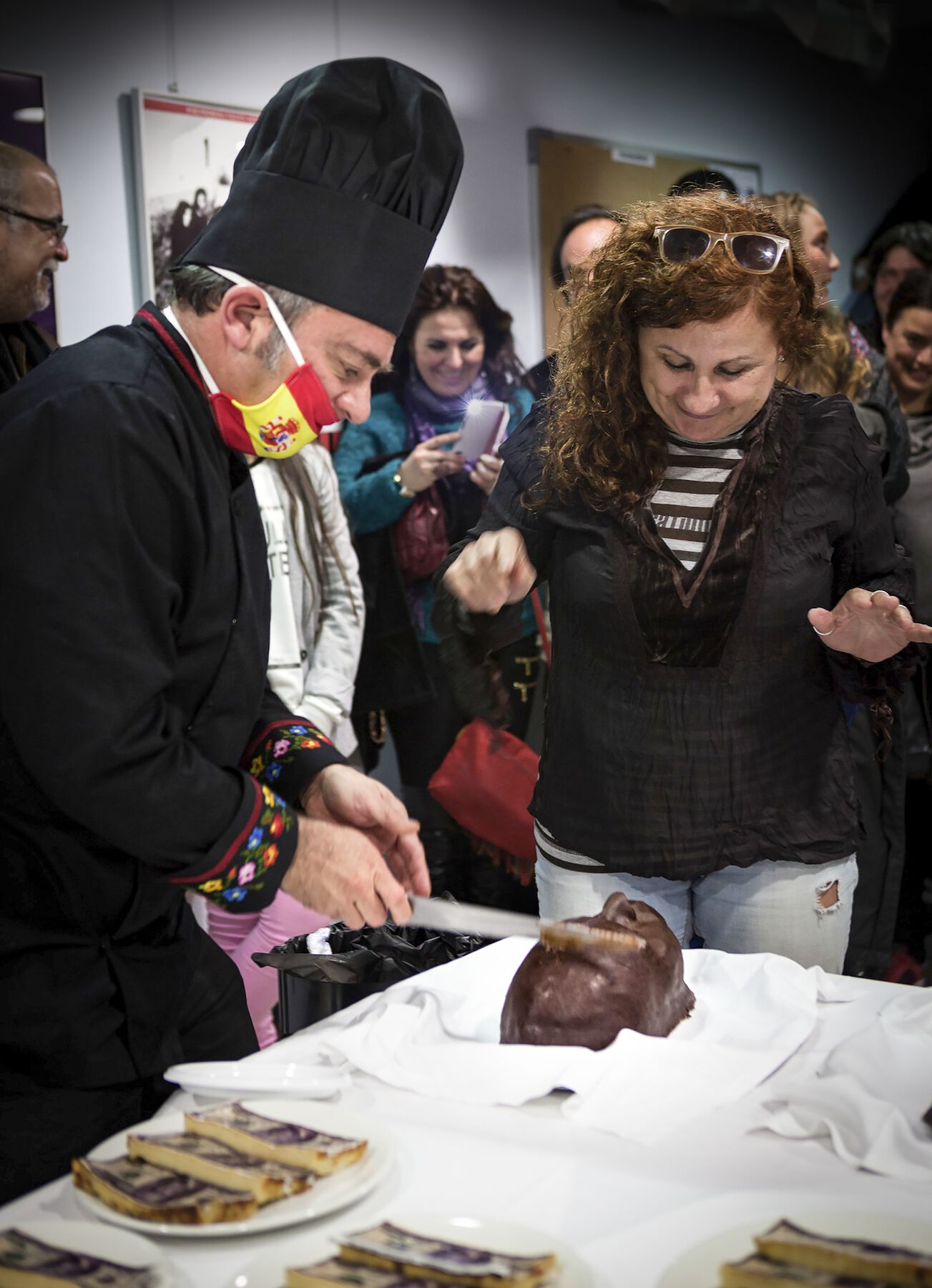 A man cutting a chocolate head sculpture starting from the forehead while a woman stands right next to him being surprised