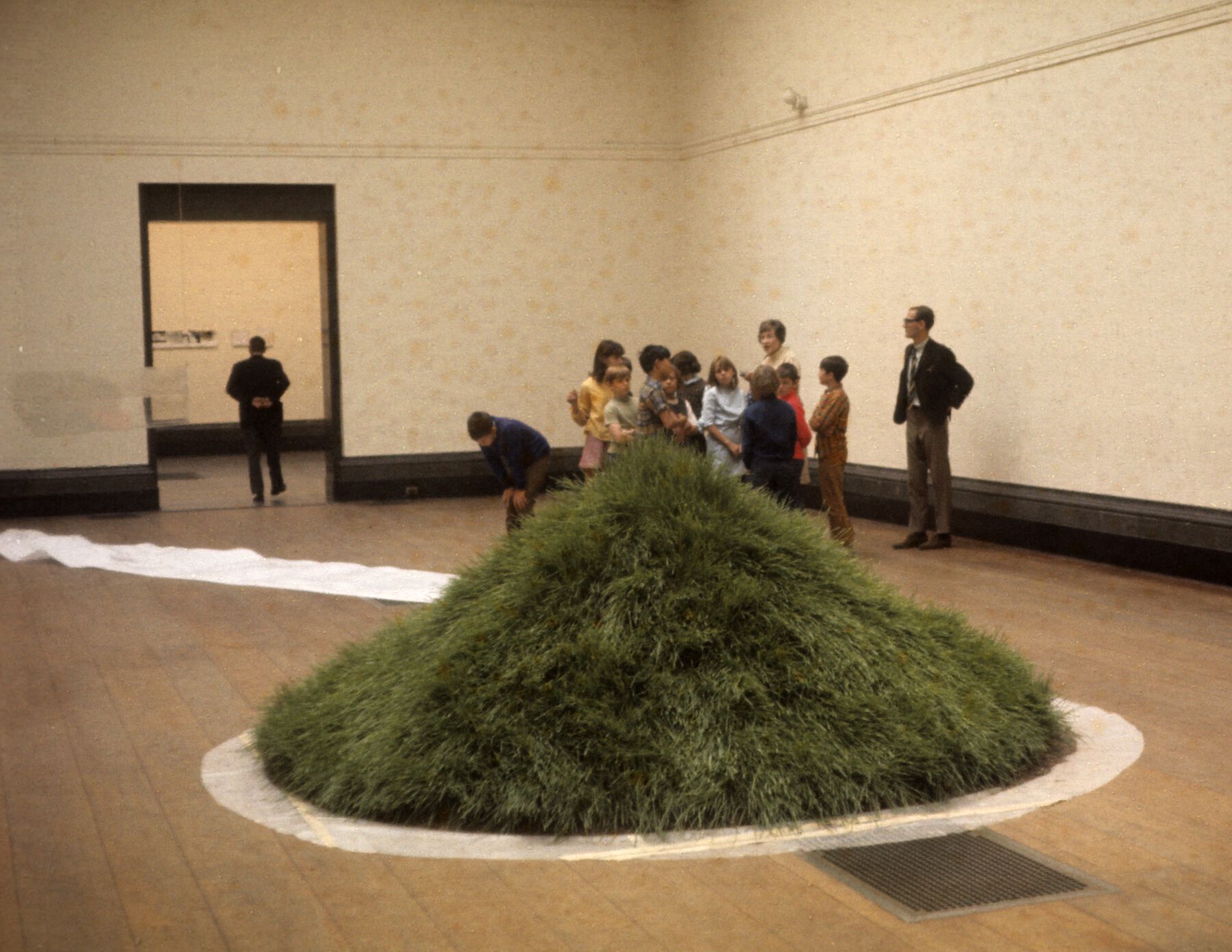 A grass mound displayed in a middle of a gallery while figures in the back inspect it from a distance