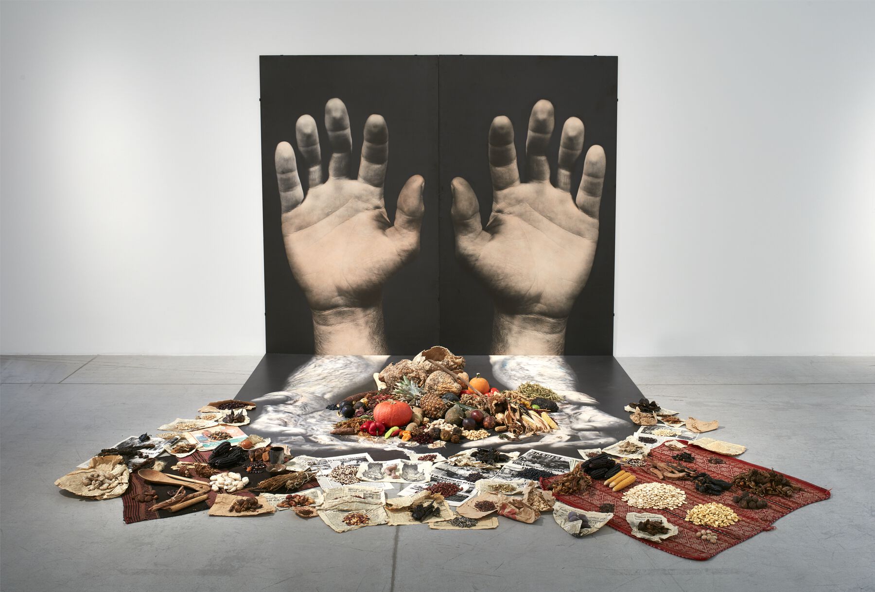 Two gigantic black and white photos of hands pasted on the wall, with food on top of tapestry and newspapers scattered in front of the photograph. 