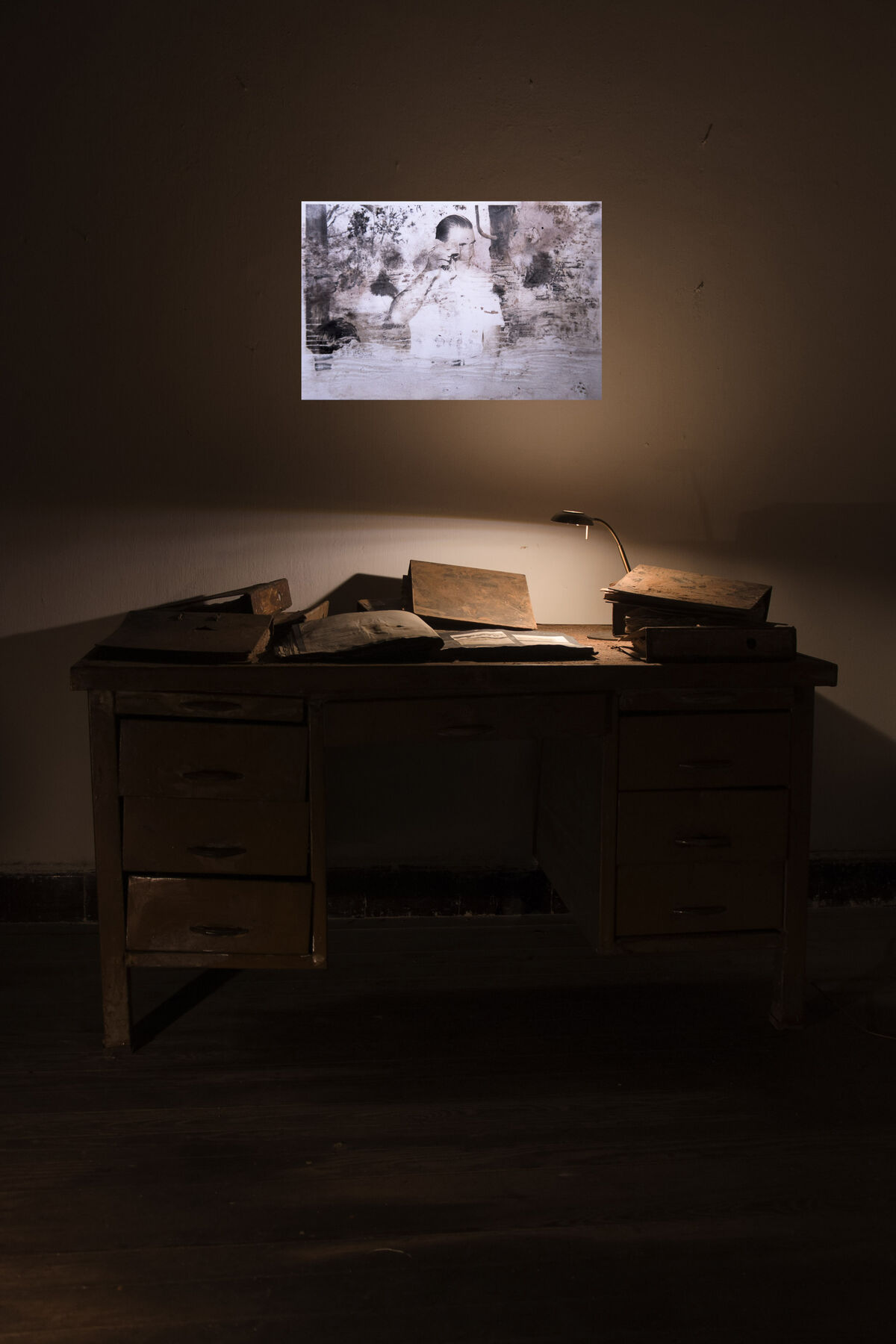 A desk with books, being lit by a lightly dimmed lamp with a black and white photograph being projected on the wall