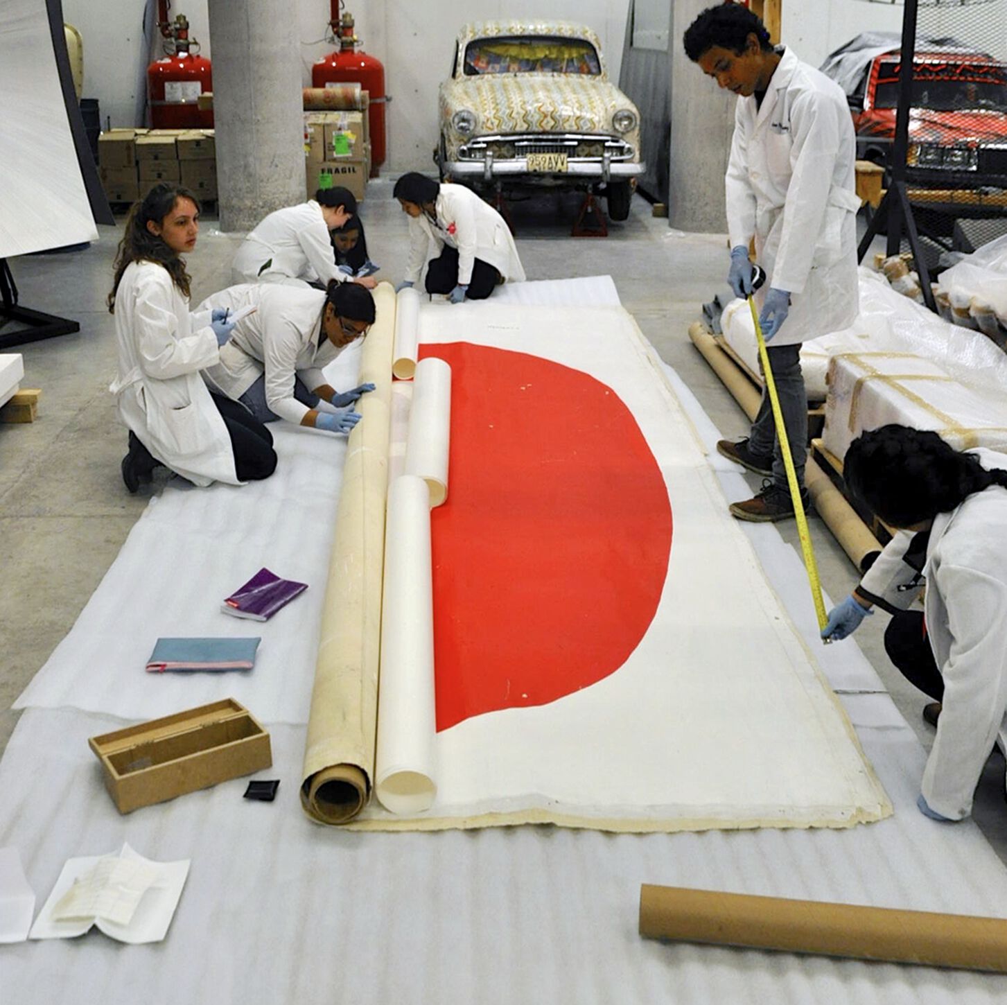 Students in lab coats measure, take notes on, and roll up, a poster adorned with a large red semicircle