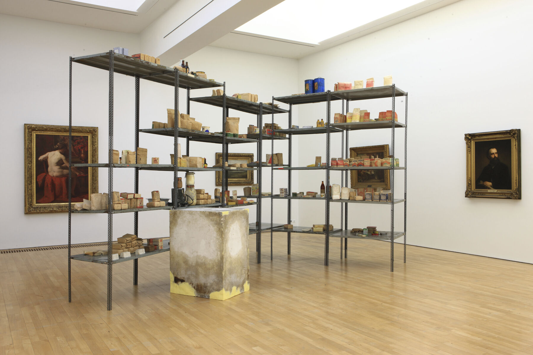 Metal shelves holding wooden objects; plaster block in front, surrounded by framed paintings in corner of room