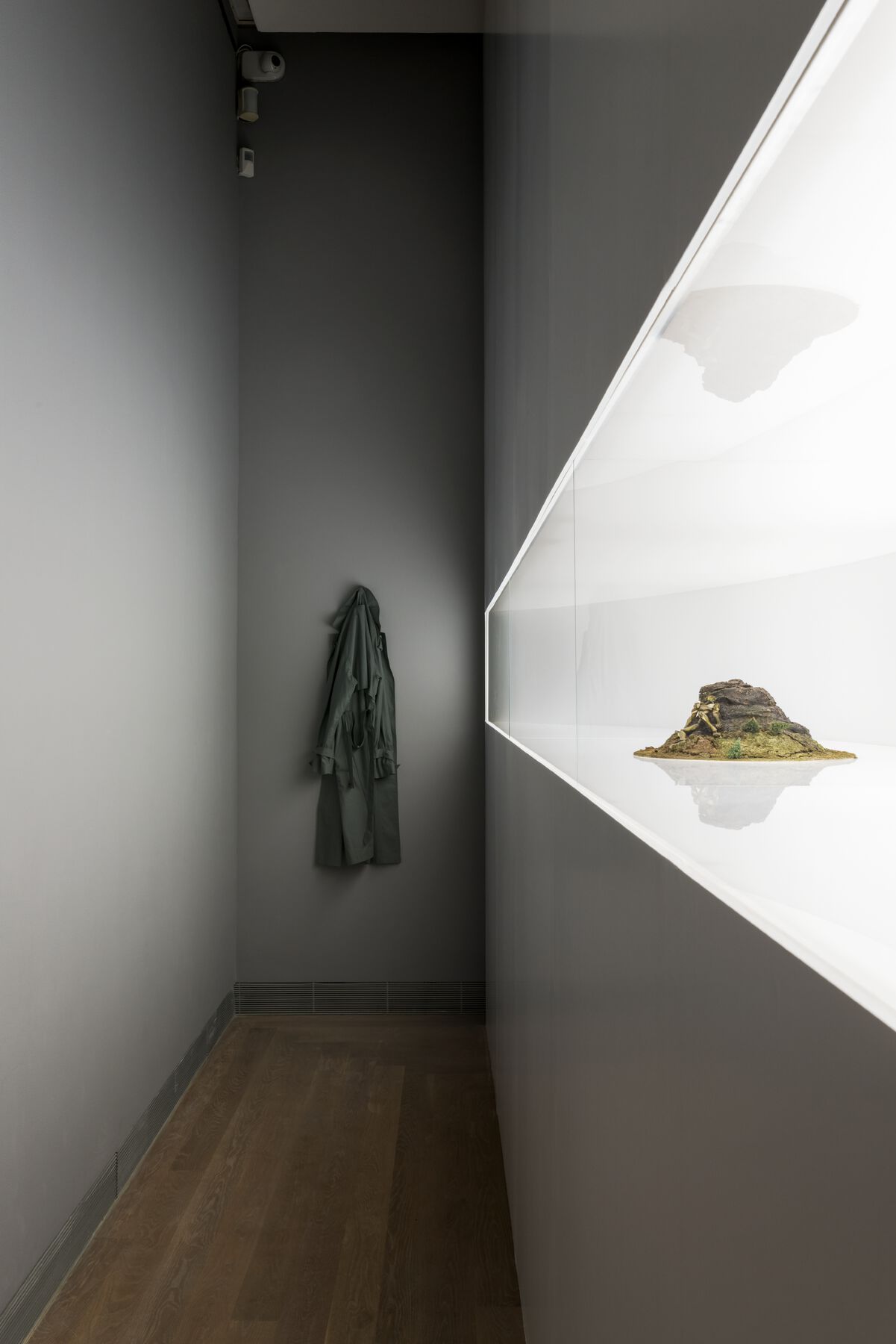 Model inside illuminated glass case in right wall of narrow, dark hallway; at hallway’s end is a hanging coat