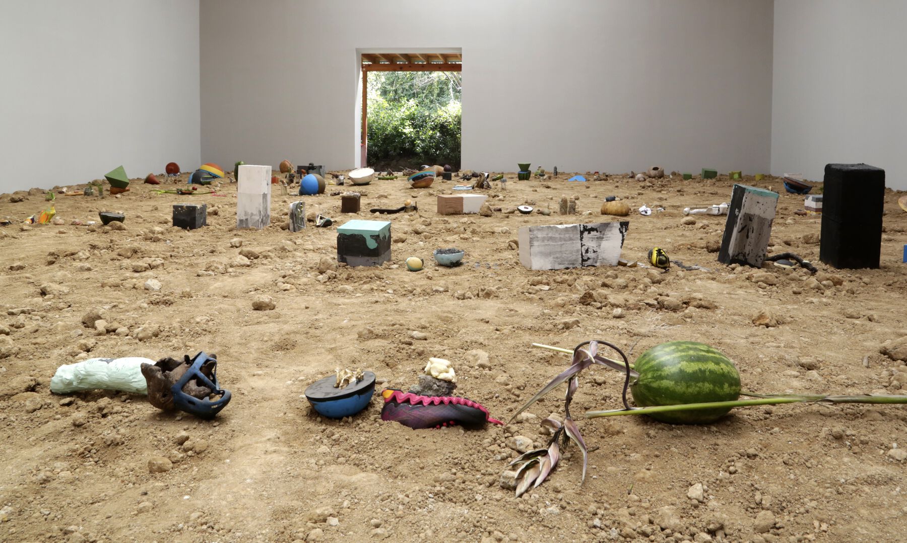 Various objects, including watermelon, plaster blocks, and pottery, buried in sand; the room has a sunlit doorway at other end
