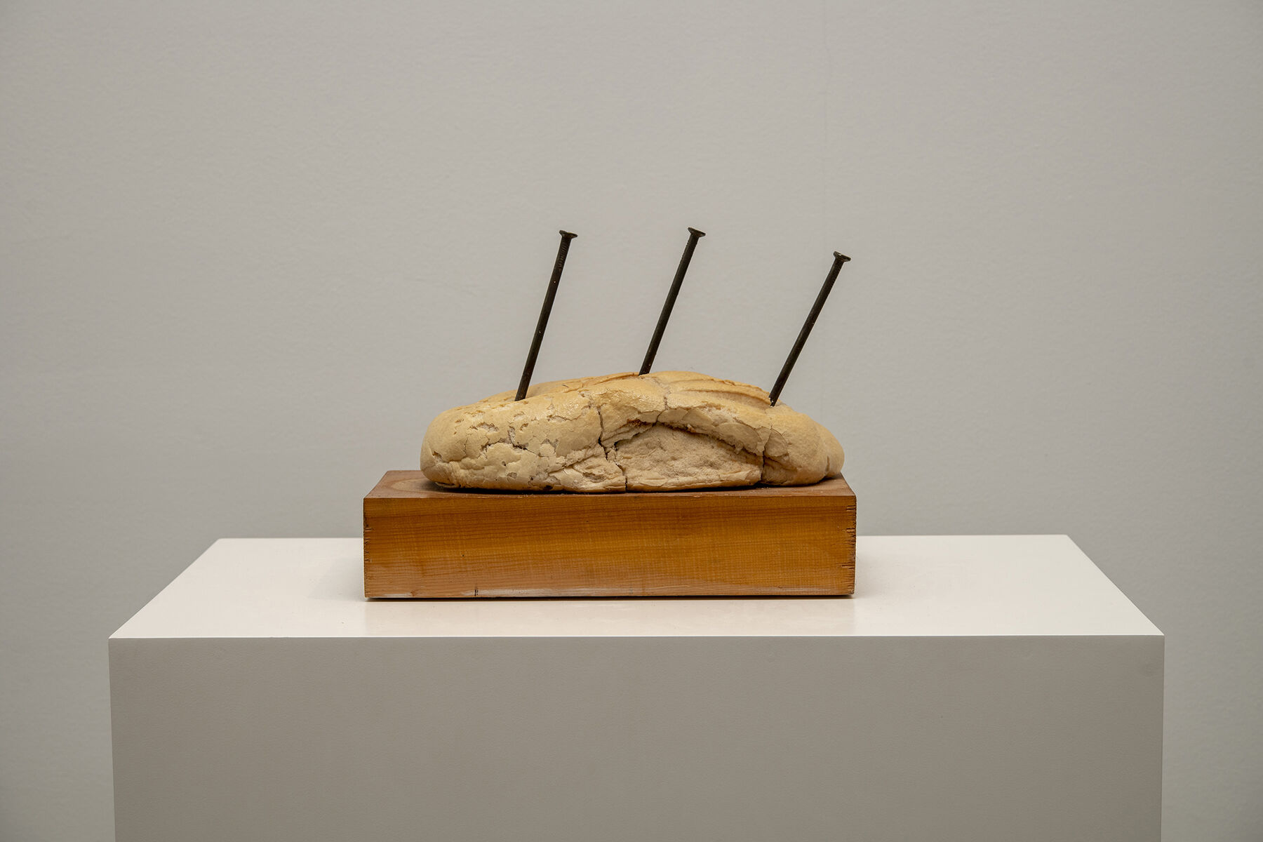 Three long black nails stuck equidistantly into crumbling bread placed upon wooden slab (atop plinth)