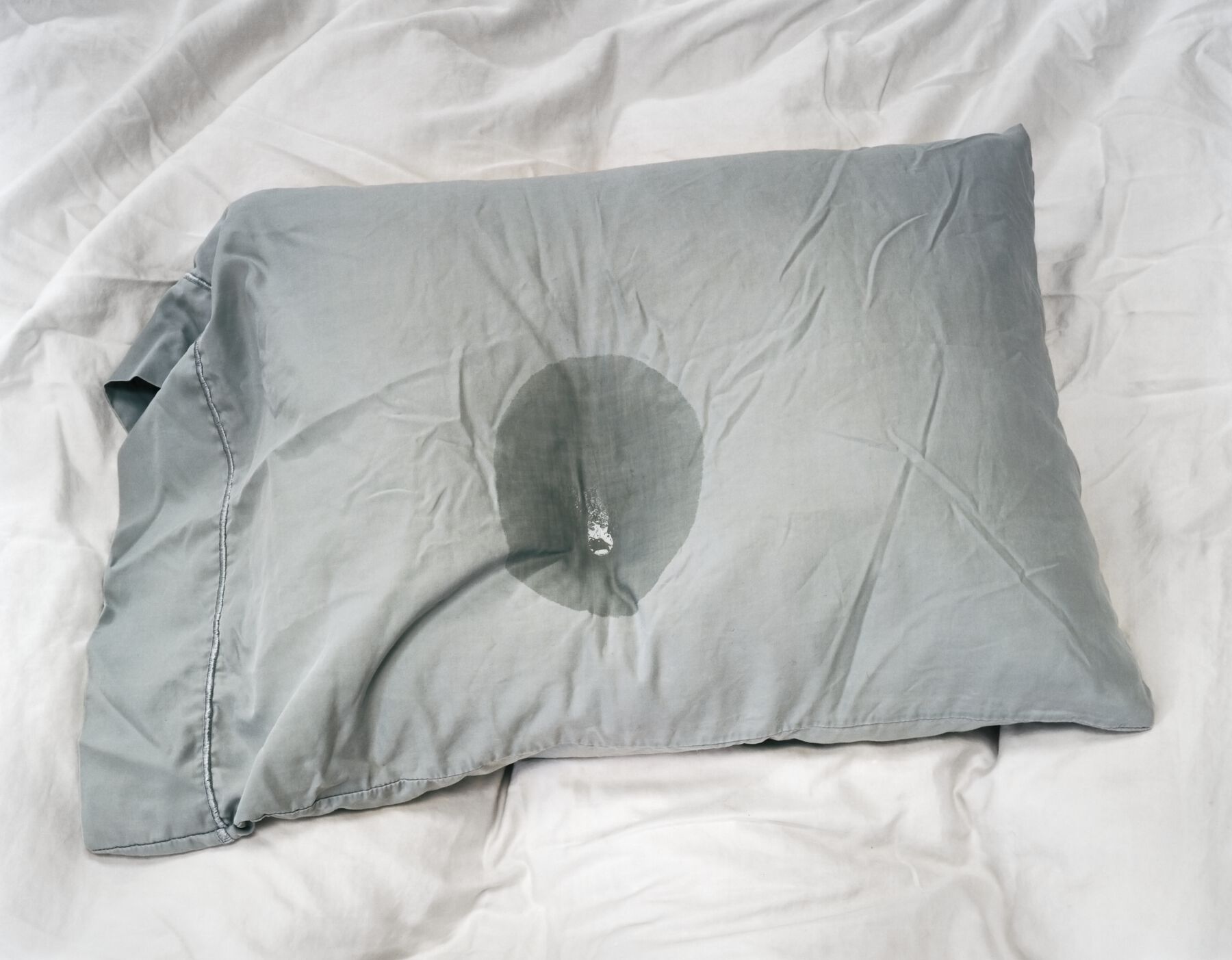 Rumpled blue-grey pillow with liquid stain (and transparent liquid pooled in center) on white bedsheet