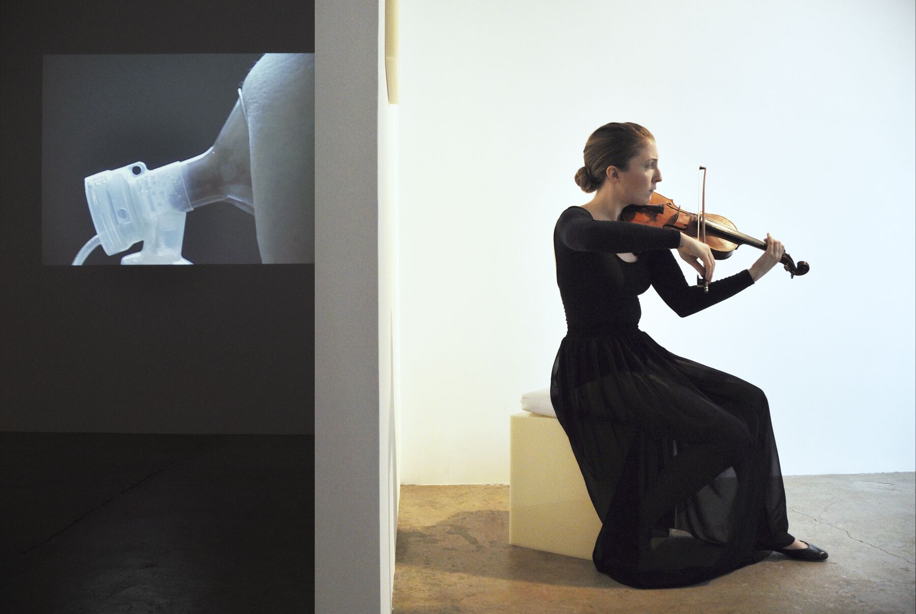 Projection of side closeup of breast being pumped (left) and musician in black dress playing violin (right) are partitioned by gallery wall
