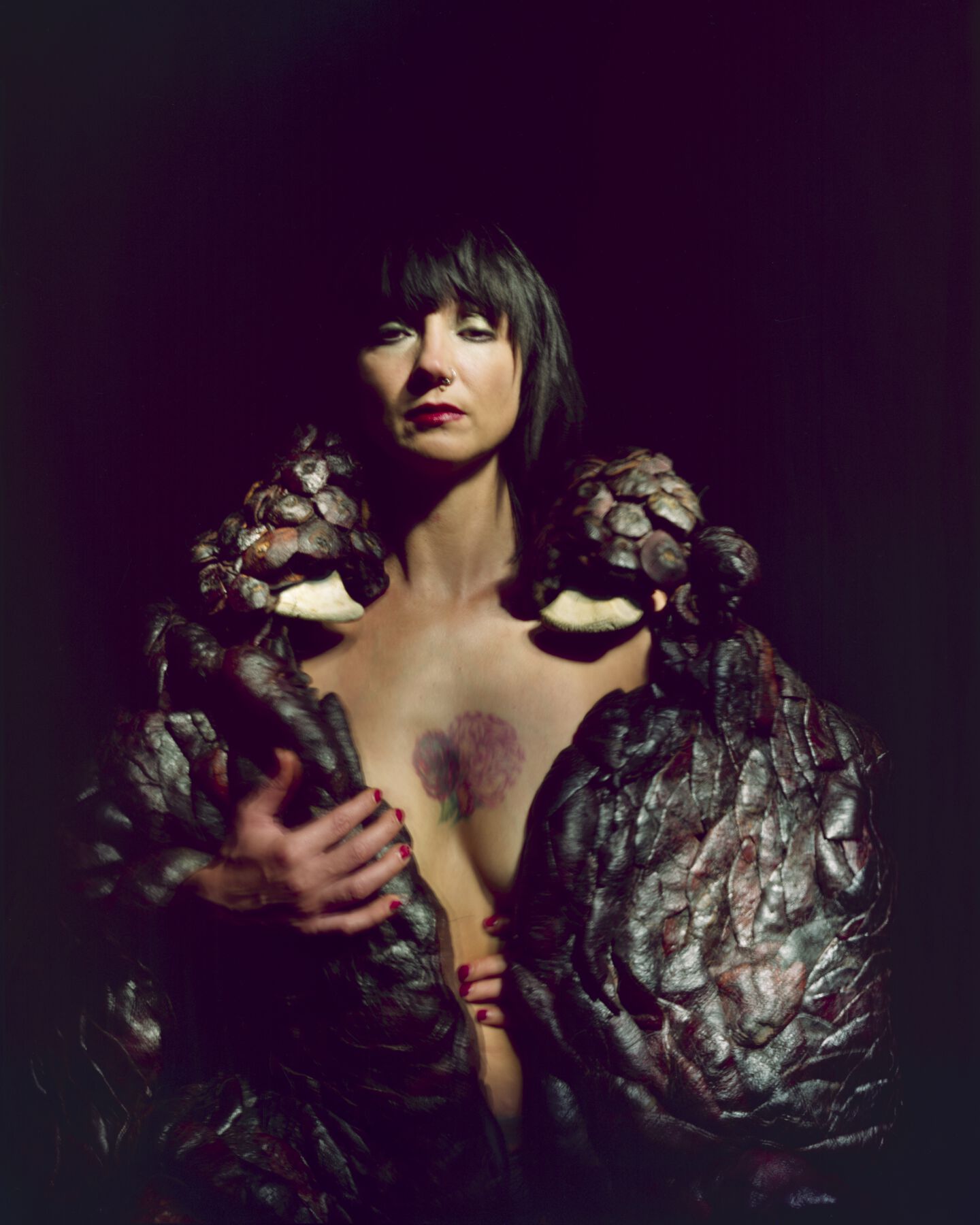 Nude artist with red lipstick and flowers tattooed on chest, wrapped in pomegranate-peel-cape; hands with red nail polish surface