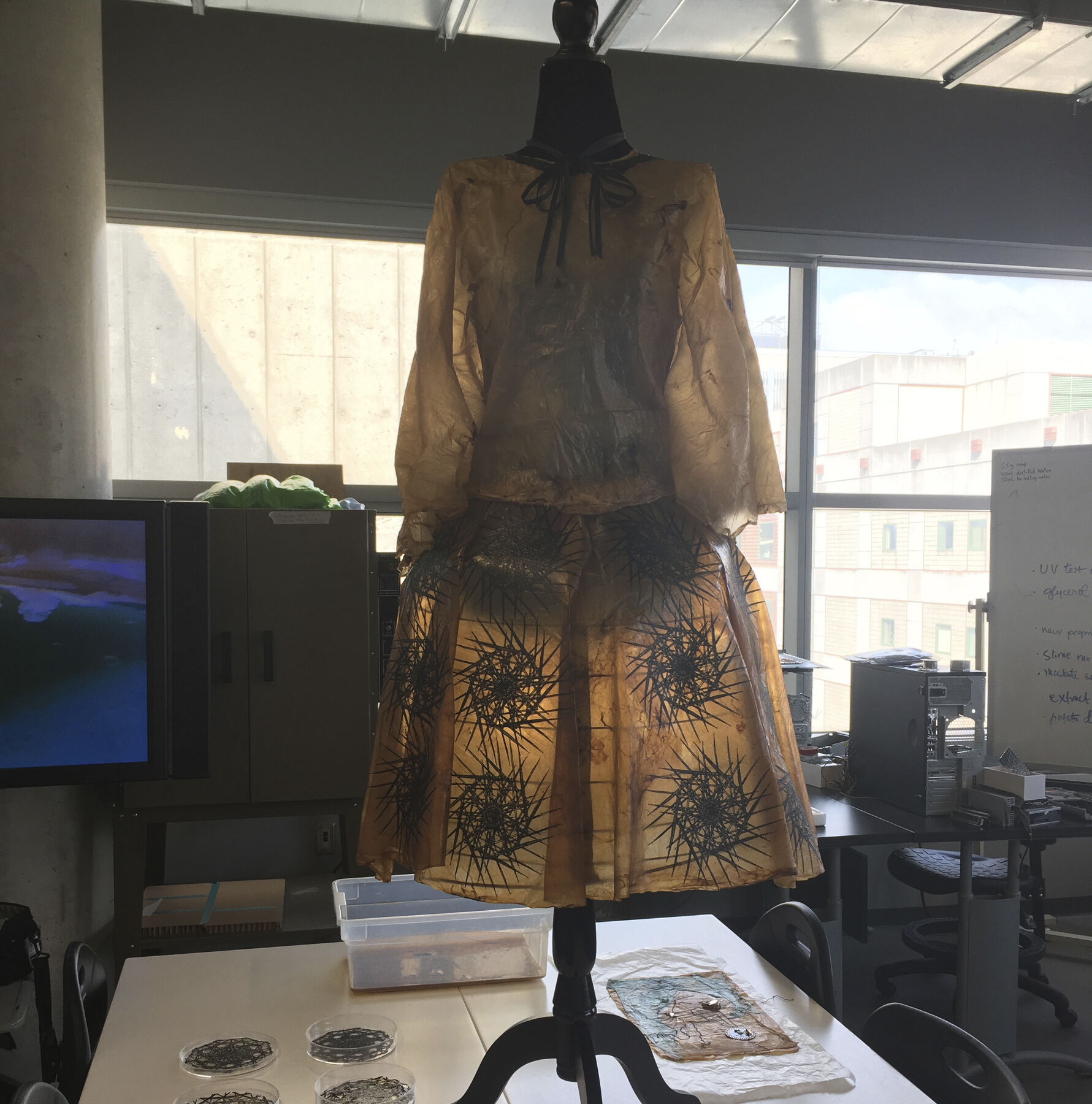 Stiff, papery translucent golden-brown dress with black geometric printed floral imagery on mannequin in sunlit fashion studio