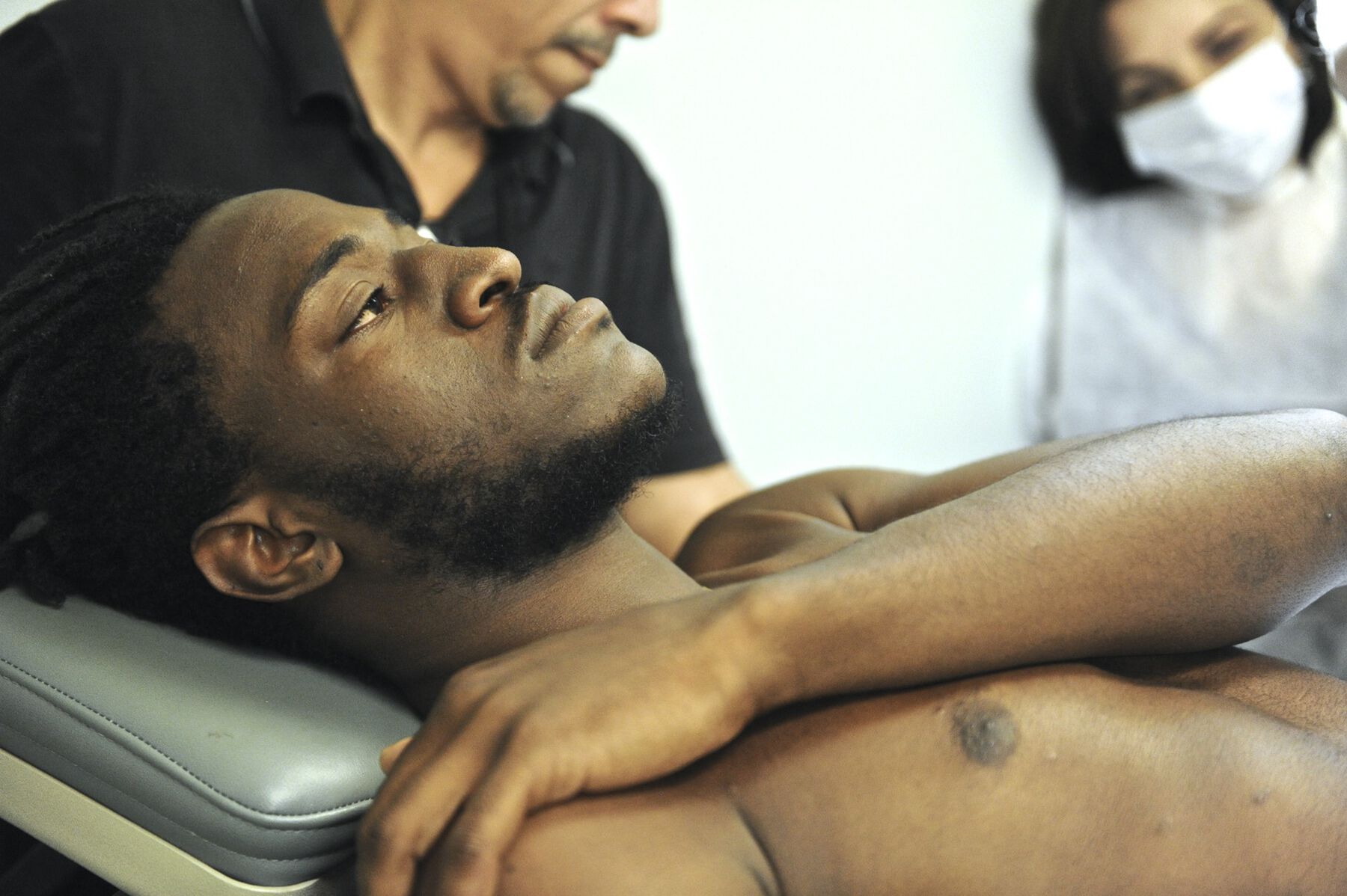 The artist laying down on a operating chair, as two figures behind him prepare for operation