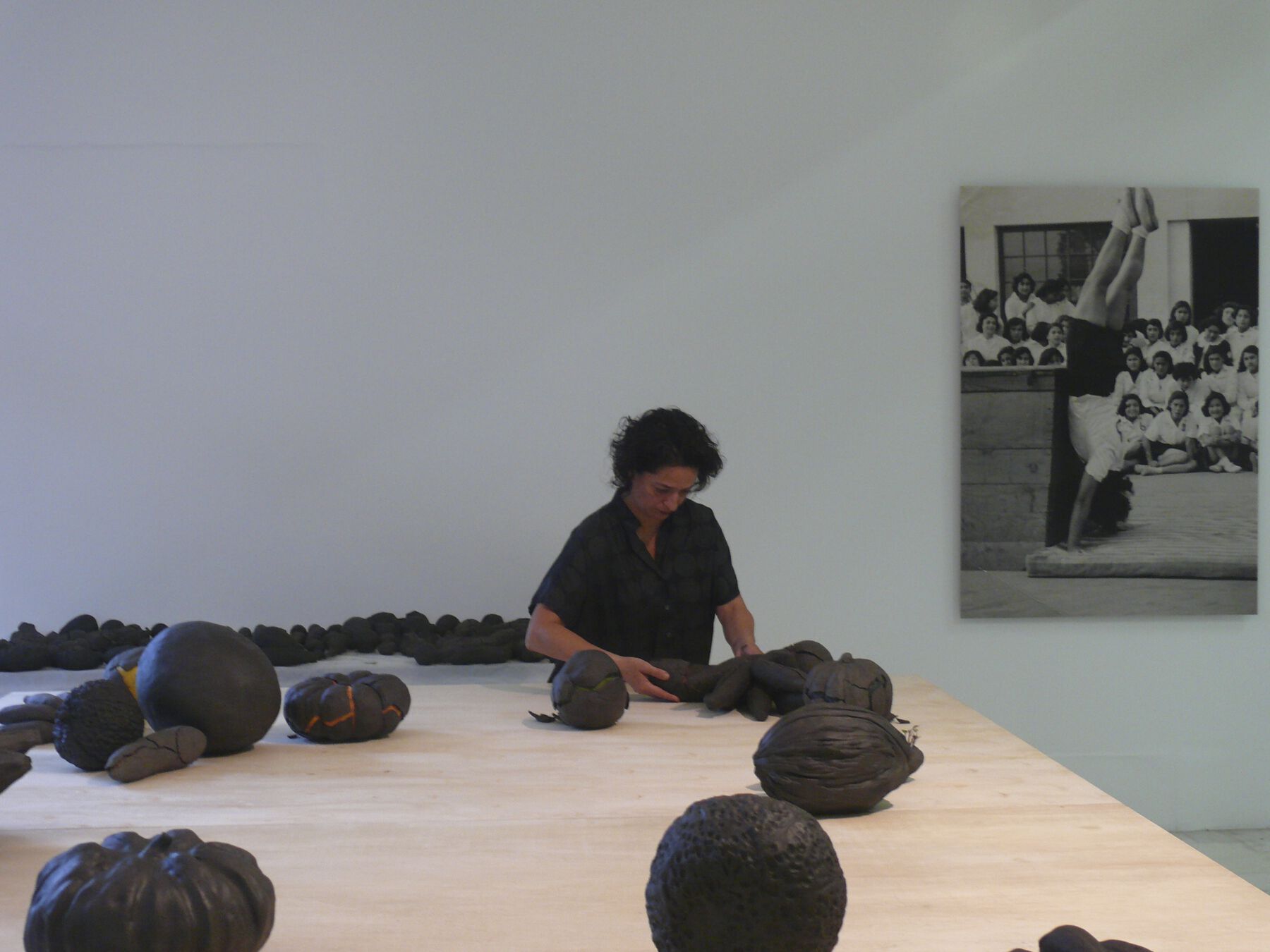 A woman standing behind a table working on covering a fruit with clay as various encased clay vegetables are scattered throughout the table