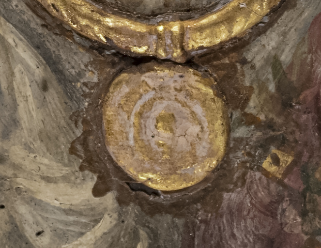 Close-up image of the pendant coin featured on a mummy portrait. There is reddish, milky crust in the scalloped outline around the gold coin. The gold paint has a nubby surface texture.