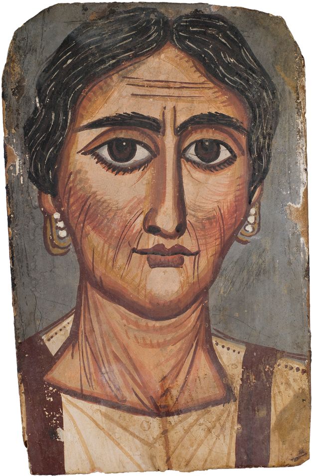 Portrait of a woman whose hair is dark with visible gray hairs. She has brown eyes and tan skin with apparent wrinkles. Her cheeks and lips have a reddish tint, and she is wearing pearl earrings. Her garment is white with brown vertical ribbon-like details on each side. The background is gray. 