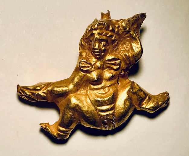 Gold amulet featuring a naked woman with her legs raised. Her left arm is raised above her, and her right arm extends out with what appears to be a club.