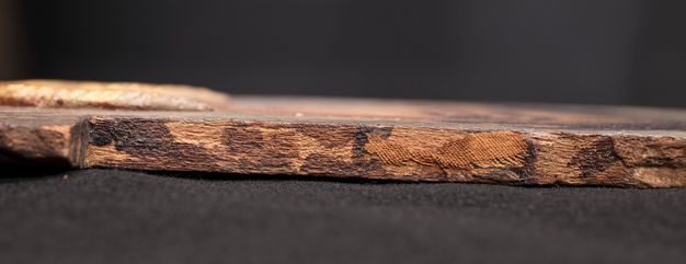 Image of a mummy portrait lying flat on a black surface as seen from the side. The left edge is shown. Gum residue and fabric fragments can be seen on the edge as dark brown stains and light brown woven remains.