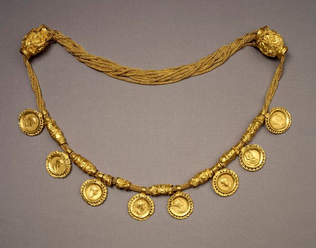 Golden necklace with pendant coins. Two large barrel-shaped charms hold together twisted, thin golden chains from which hang eight pendant coins separated by gold beads. The coins feature the profiles of individuals in relief.