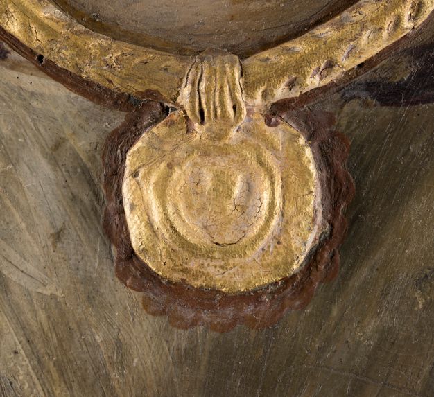 Close-up image of the pendant coin featured on a portrait. There is reddish, milky crust in the scalloped outline around the gold coin. The gold paint has a nubby surface texture.