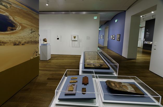 Photo of the funerary portrait installation at Harvard Art Museums. There are three glass display cases in the room, and the bust of a man's head is visible around the corner in another glass case. Two of the three cases in the foreground have portraits, one is full-body-sized while the other is a portrait of the shoulders and up of a man. In the third case, there are five small artifacts. The wall on the left is a blown-up photo of a desert and small body of water with a few shrubs.
