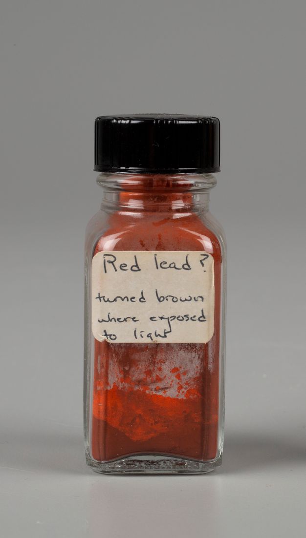 Picture of a small glass jar that is less than halfway full of a red powder. The white label on the jar reads, “Red lead question mark. turned brown where exposed to light.” The powder clinging to the inner walls of the jar is turning brown.