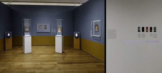 Photo of the funerary portrait installation at Harvard Art Museums. There are five portraits on display, two are in glass cases on a pedestal, two share a glass case on the back wall, and the fifth has its own case on the right wall.