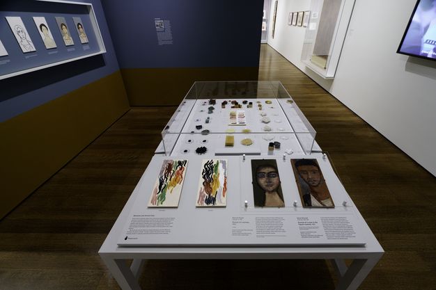 Photo of the funerary portrait installation at Harvard Art Museums. There are student copies of the portraits displayed on the left wall. A center table displays materials for funerary portraits with two touchable portraits and two canvases with paint squiggles for a tactile experience.