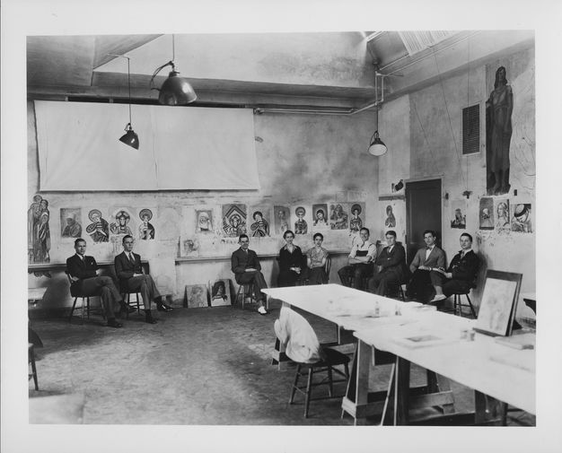 Historic black and white photo of a group of people sitting in a room along the wall. There is a long table in the center of the room. Portraits hang on the walls behind the people.