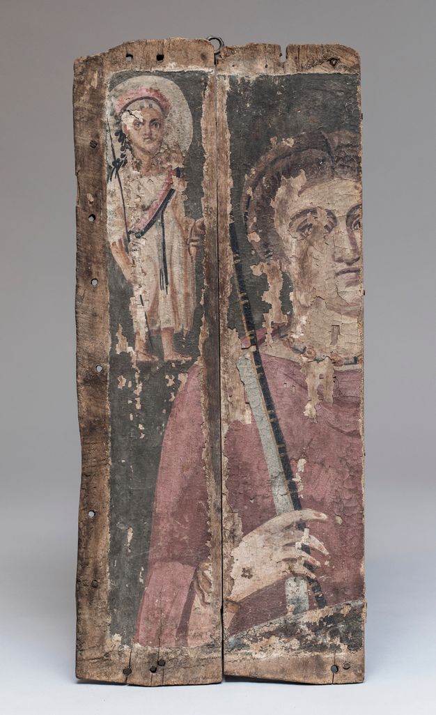 Fragmented votive panel painting that depicts the goddess Nemesis, who takes up most of the painting, and the god Harpocrates, who is small and appears in the upper left corner. Nemesis holds a cubit, or measuring stick, in her right hand and wears a pinkish tunic. Harpocrates has a large white halo behind his head, and he has a garland and situla. The paint is chipped in several places.