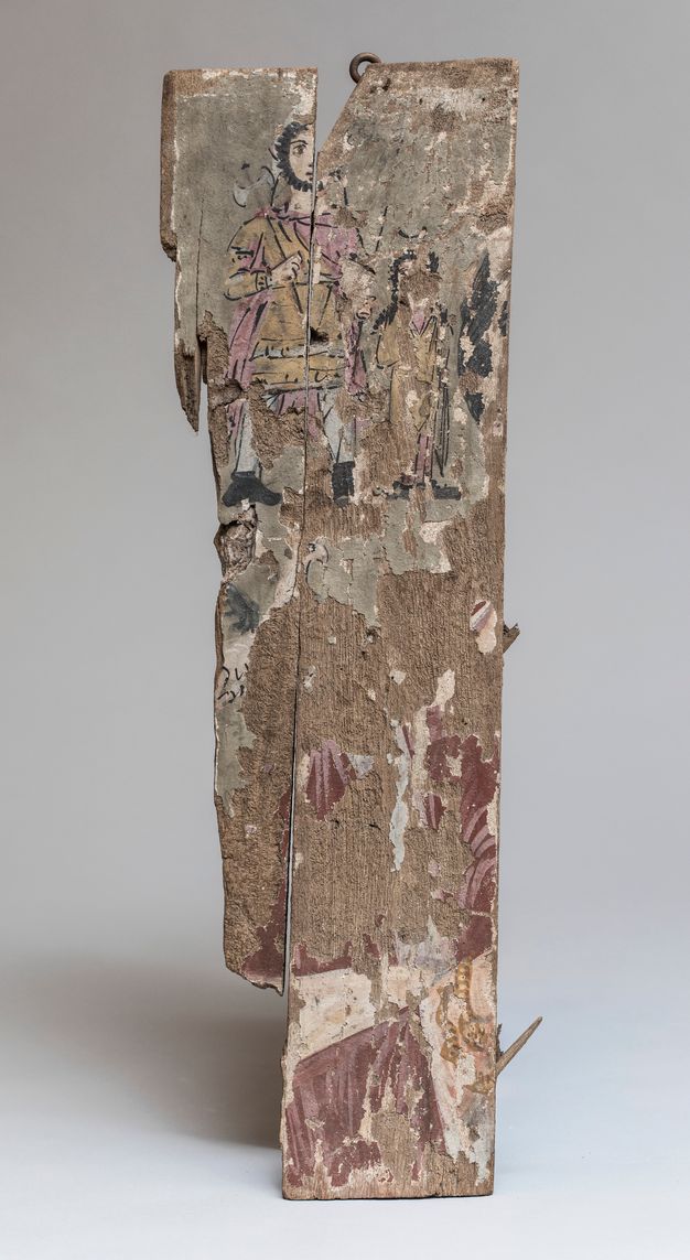 Fragmented votive panel painting rendering a female bust, a winged griffin, and two minor divinities. Most of the paint has chipped away, but the two minor divinities are the most visible of what remains. They are in the upper half of the painting, and the one on the left is taller than the other. They both have short hair and beards, and the taller one holds an ax. They both wear tunics of faded yellow and pink. The female bust is recognizable by the right shoulder still visible at the bottom right. A gray clavus is visible on the tunic, and a whitish mantle is arranged across the figure’s abdomen. Her sleeve is decorated with a white fringe at the opening and she wears a gold snakelike bracelet with two twisted rows.