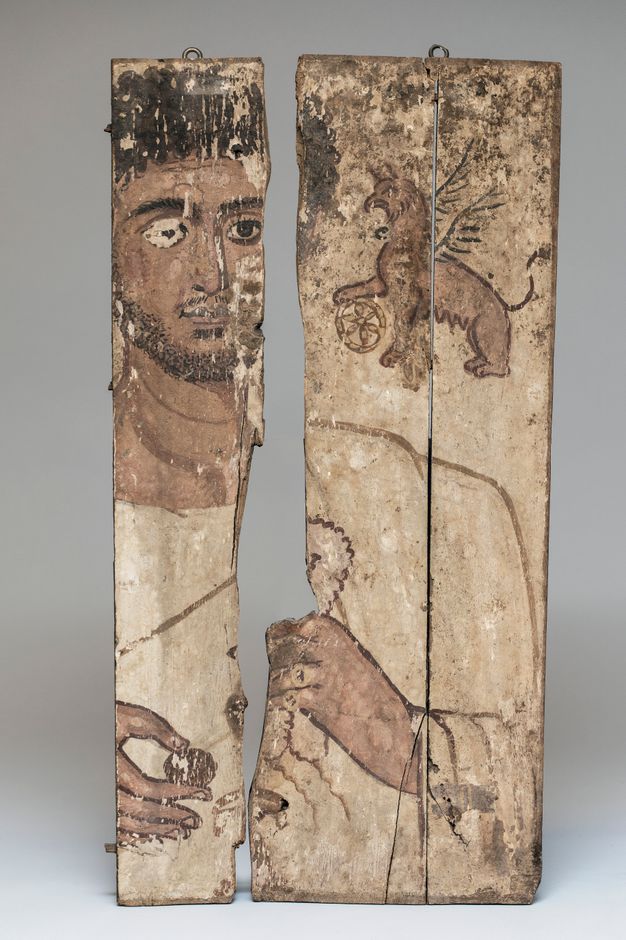 Fragmented portrait of a man and a griffin with the wheel of Nemesis. There is a missing piece in one of the boards, so that the portrait is presented in two sections with the man's head on the left and his left shoulder and the griffin on the right. The griffin hovers above the man's left shoulder with a paw on the wheel of Nemesis. The man wears a white tunic and white mantle. He holds one object in his left fist and another pinched between the thumb and forefinger of his right hand.