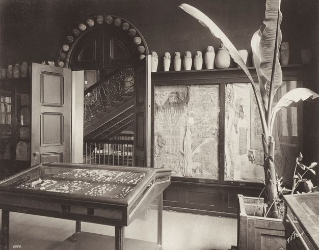 Black and white photograph showing a gallery at the Copley Square Museum. There is a display case in the center of the room in front of the doorway. To the right, there is a glass display with two shrouds.