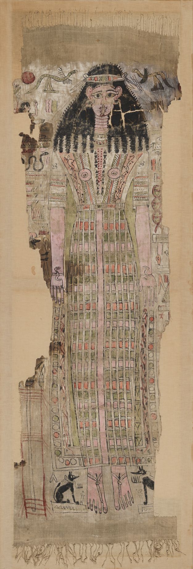 Photo of the Hathor shroud in color. The shroud has tasseled fringe at the top and bottom edges and is painted in red, orange, green, pink, and black on a white ground. It bears a full-length figure of the deity Hathor, whose long hair ends in thick curls, wearing a long, striped headband and an embellished tunic under a feathered sheath dress with wide straps decorated with vines. An undecorated column runs down the center of the dress from waist to ankles. The tunic has decorated sleeves and the breasts are covered with rosettes. The figure wears various adornments, including a jeweled circlet; large hoop earrings; a long, beaded amulet; decorated pectoral; snake bracelets on either wrist; rings on both hands; and thong sandals. She is surrounded by hieroglyphs and mythological creatures, two Anubis jackals flanking her feet and mythical serpents in the top registers, and various architectural and vegetal elements. There are considerable losses to the textile at the sides and to the paint and ground, as well as considerable staining across the face and proper right arm of the central figure.