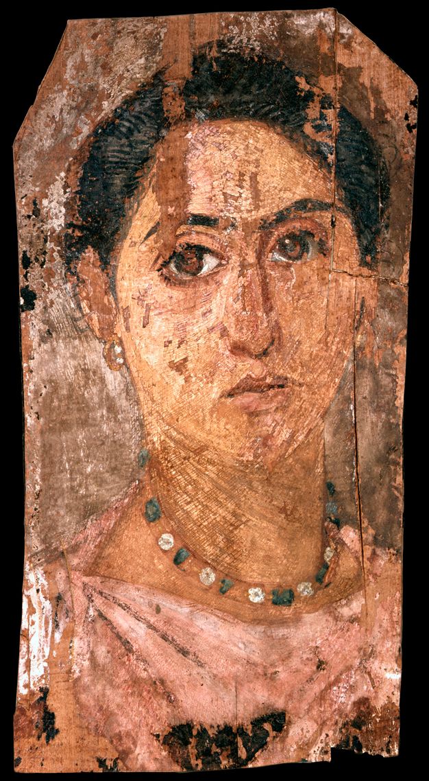 Portrait of a woman with dark brown hair and brown eyes. Her hair sits on top of her head, and she is shown wearing a pink tunic, gold earrings, and a green and white necklace.